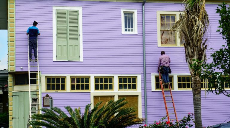 Two painters working on ladders to paint the exterior of a wooden house in a bold lavender color with cream and yellow trim accents.