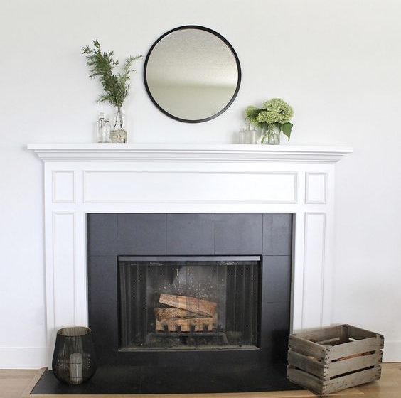 Modern fireplace with a white mantel, black tile surround, and minimalist decor featuring a round mirror and greenery.