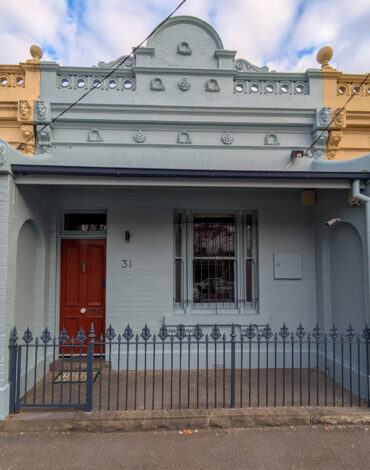 Single-fronted Victorian terrace on Kay Street, Carlton, repainted in pale blue-grey with a red front door and black palisade fence