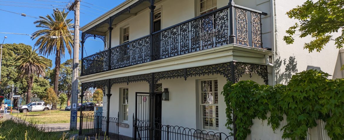 Victorian-style house with ornate ironwork balcony, lush greenery, and palm trees.