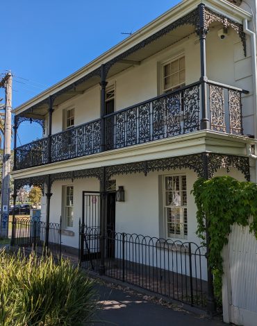 Victorian-style house with ornate ironwork balcony, lush greenery, and palm trees.