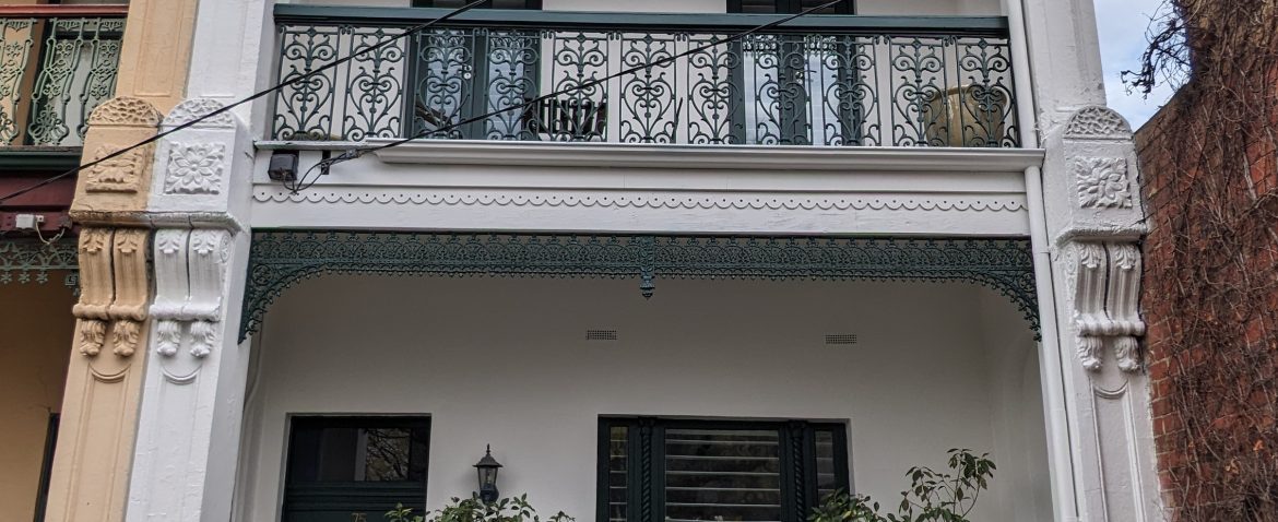 Facade of a traditional two-story building with intricate ironwork and greenery in the front garden.