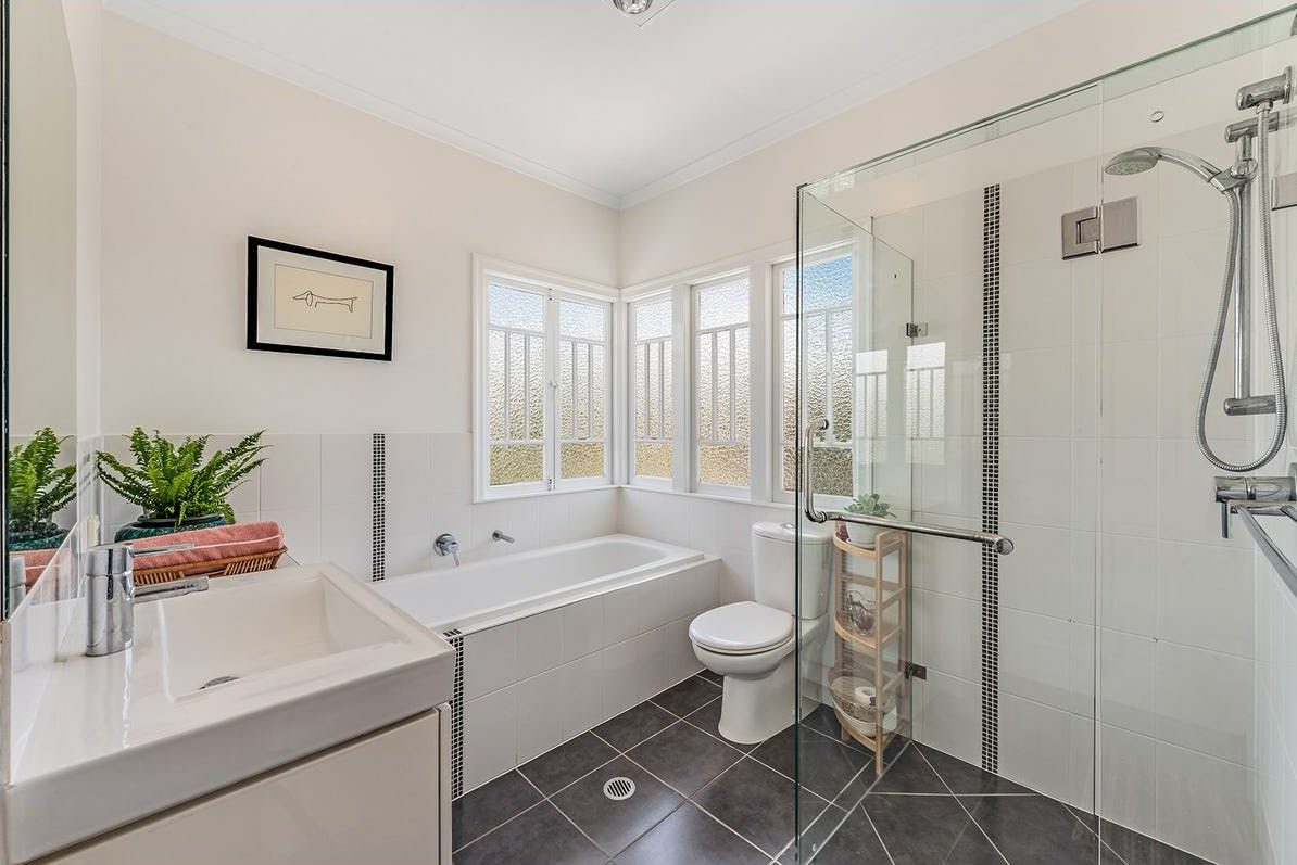 A bright and modern bathroom featuring a bathtub, glass-enclosed shower, white vanity, and large frosted windows allowing natural light. The space is decorated with a framed artwork, potted plants, and a small wooden shelving unit.