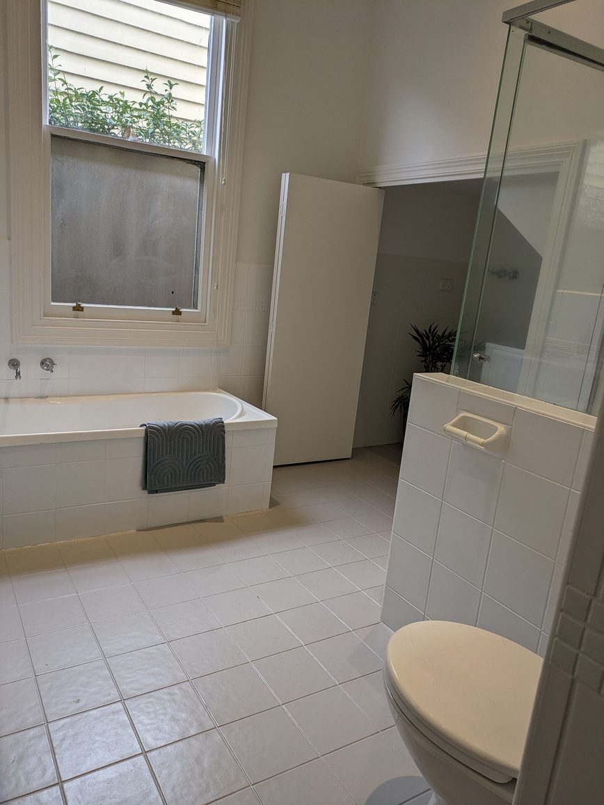 Simple white bathroom with a bathtub, towel hanging, glass shower, toilet, and a window with a view of greenery outside.