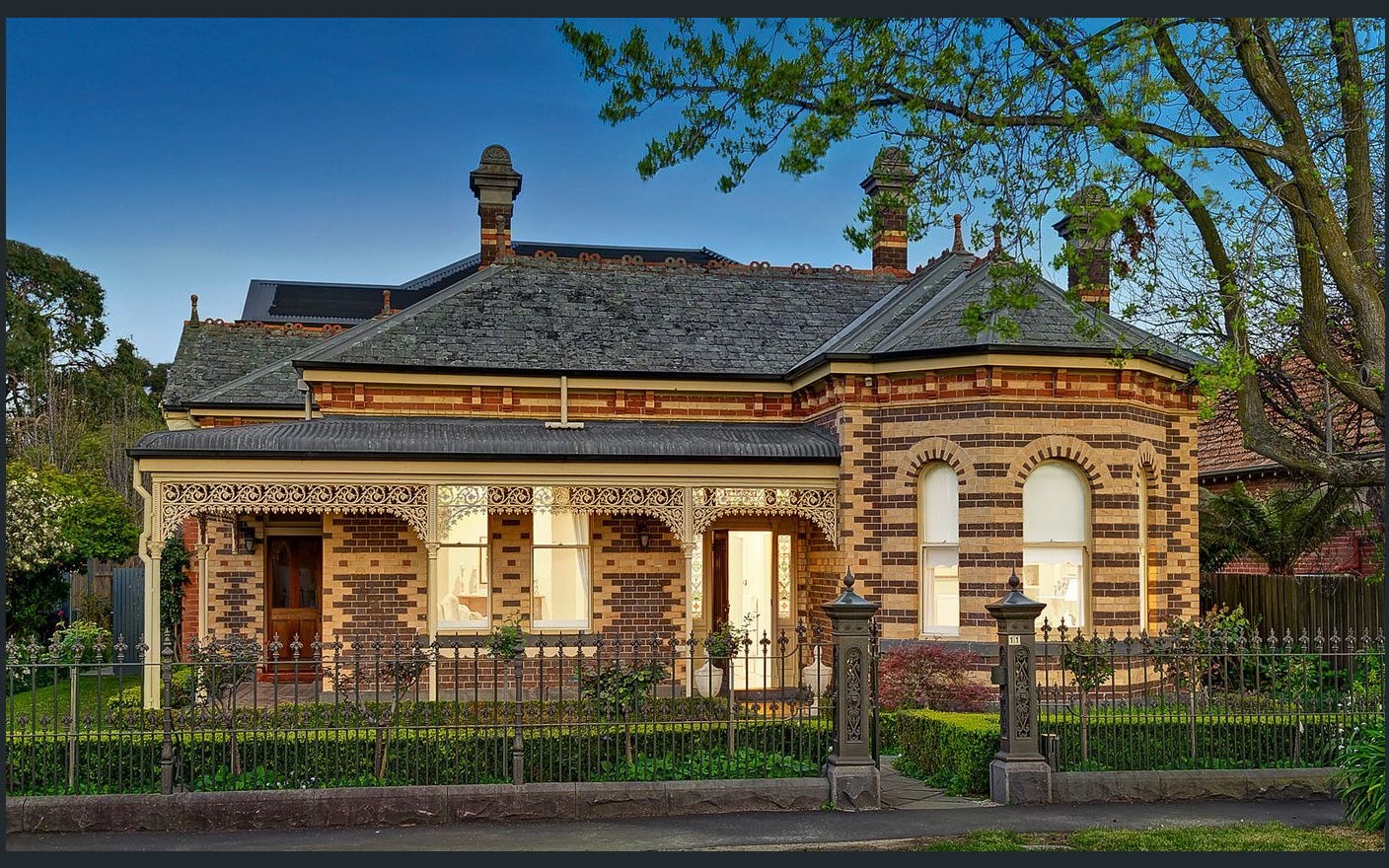 Heritage-style house with brick facade, decorative ironwork, and beautiful garden, captured at dusk.