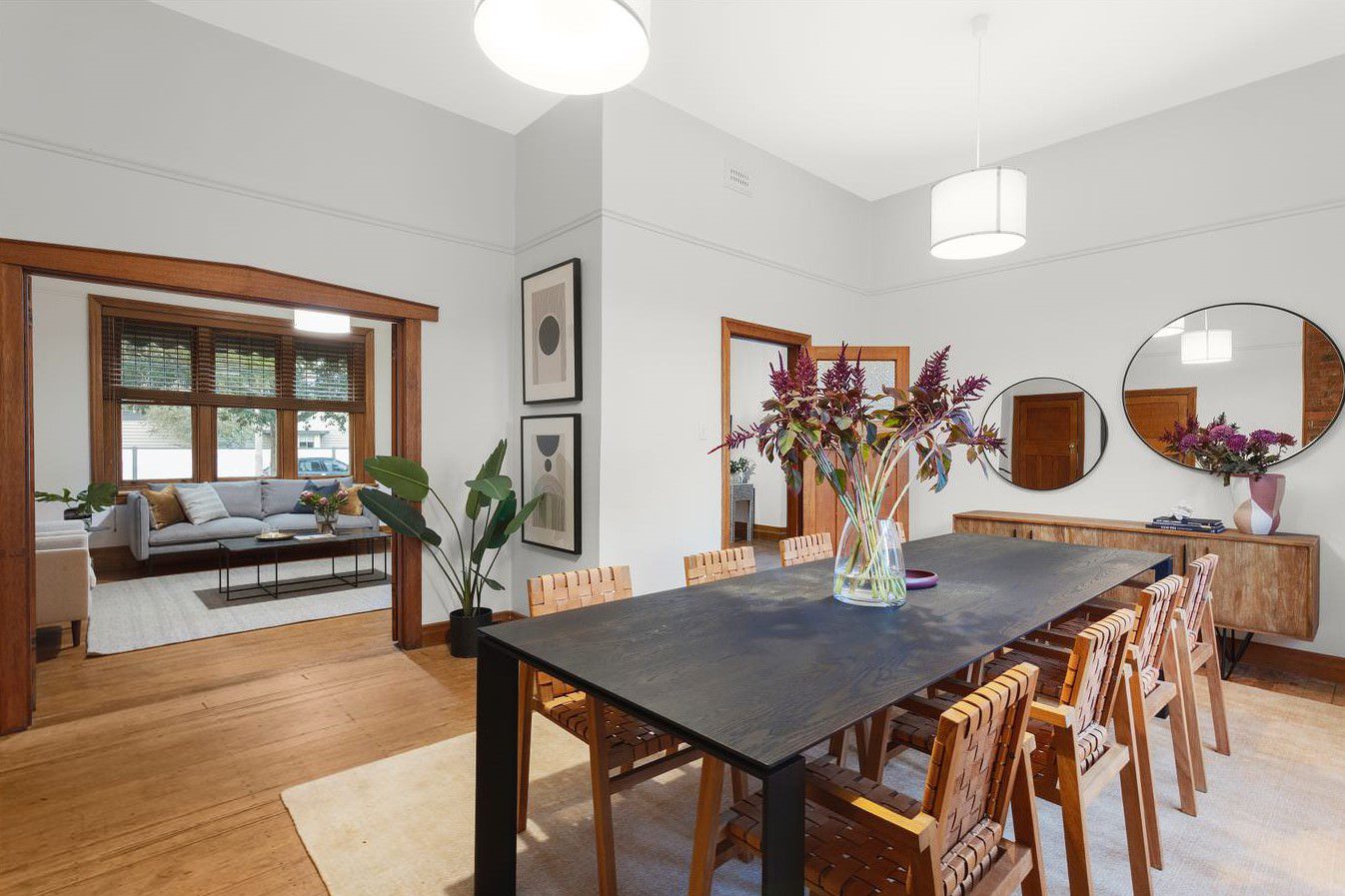 A stylish dining room with a black wooden table, woven leather chairs, and a sideboard with decorative mirrors. The space is adorned with modern artwork, plants, and a floral centerpiece, leading into a cozy living room with large windows and a neutral-toned sofa.