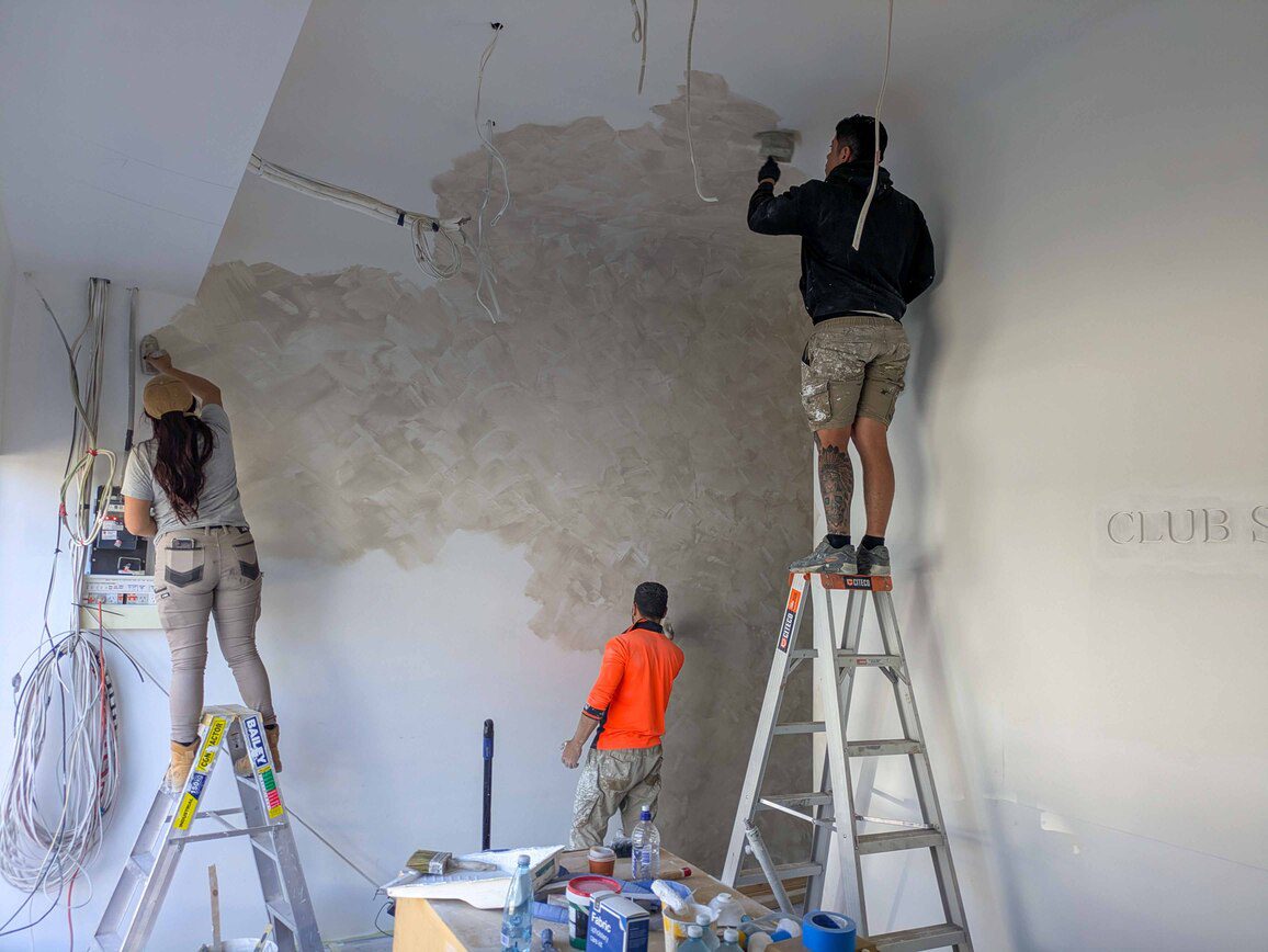 Three painters working on a textured feature wall, with two standing on ladders and one on the ground, carefully applying a decorative finish in a modern interior space.